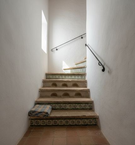 Narrow whitewashed stairwell with patterned tiled risers, worn stone treads, a black iron handrail and a folded blue striped cloth