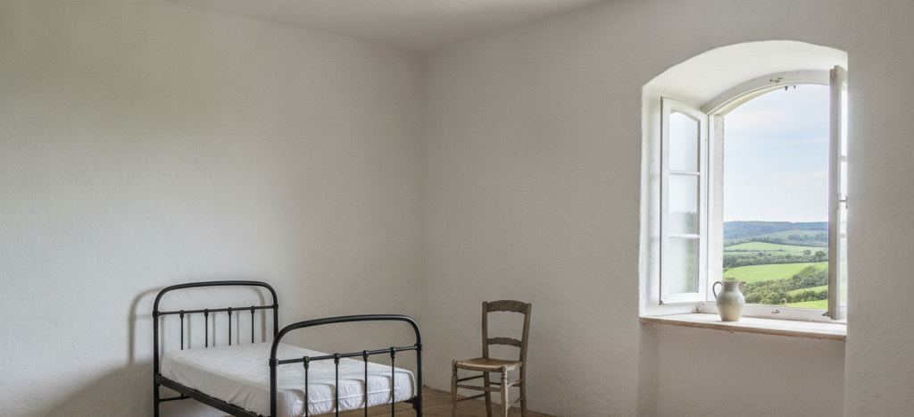 Bare whitewashed room with an iron bed frame, a worn wooden chair, and an arched window open over green hills