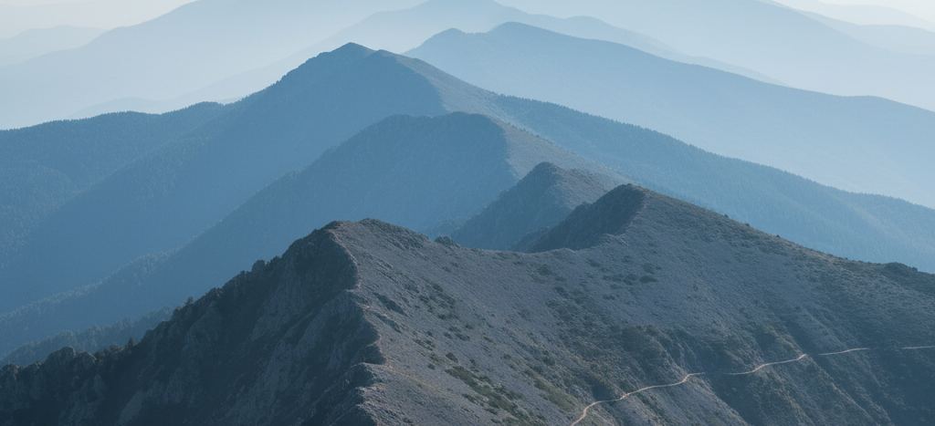 A bare rocky ridge crest with a thin footpath traversing it, forested slopes below and pale blue ridgelines fading back into haze