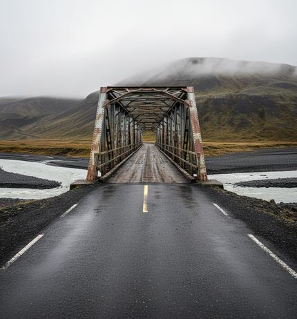 Wet two lane road narrowing into a rusted single lane truss bridge over pale braided river channels, mossy hill under low cloud