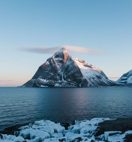 A snow streaked dark peak across a still fjord, a thin cloud snagged at its summit, ice crusted rocks along the near shore
