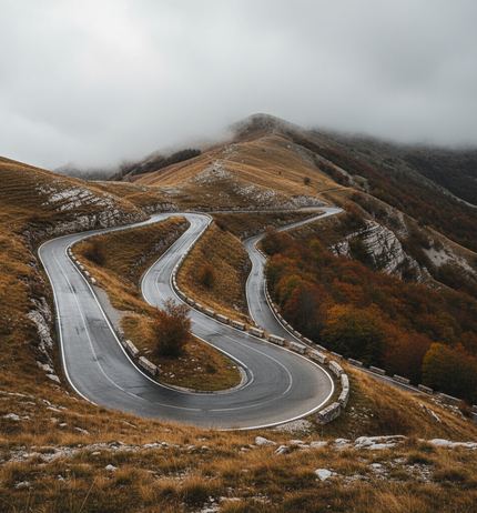 A mountain road doubling back in tight hairpins over a grassy ridge, white barrier posts on the outer bends, rust autumn scrub, cloud on the summit