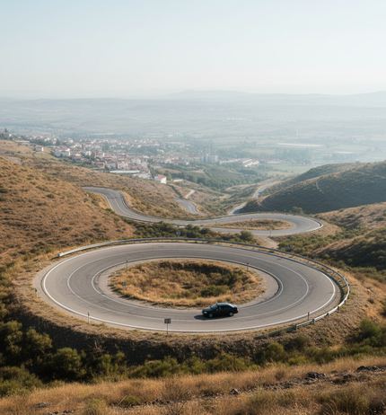 A road looping in a full circle down a dry golden hillside, a single dark car on the curve, hazy town and valley below