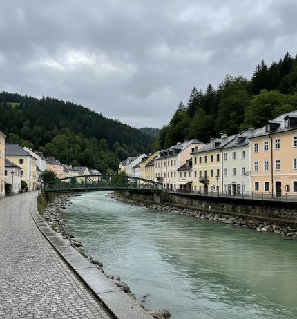 A fast pale green river between a wet cobbled quay and cream and yellow townhouses, a green steel bridge beyond, wooded hills under grey cloud
